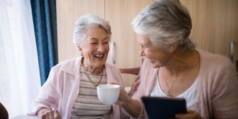 Two Elderly Women Laughing Over Coffee at Assisted Living Facility